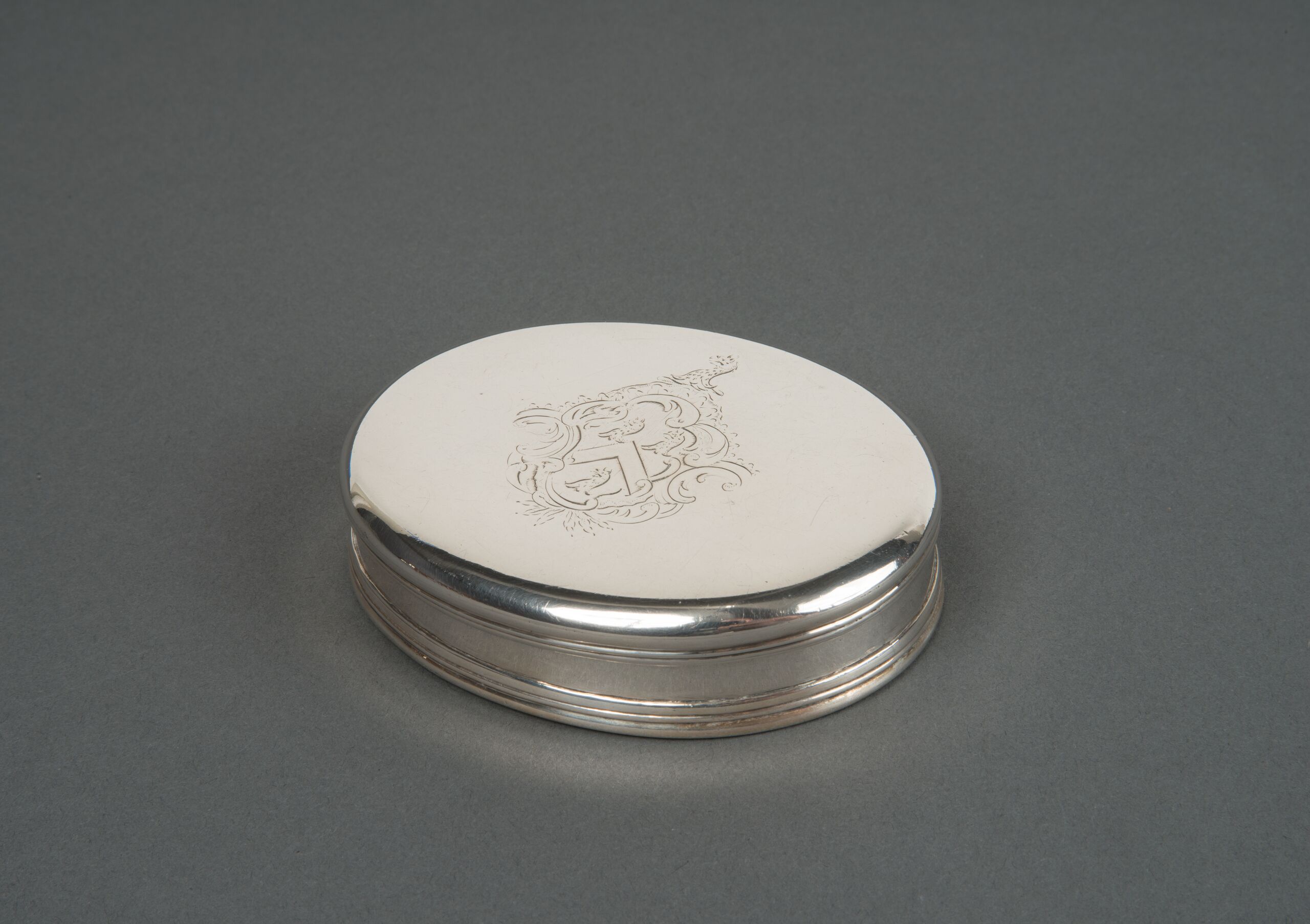Small oval tobacco box made of silver. The relatively plain, closed box features a Rococo engraving of a coat of arms at the center of the lid.
