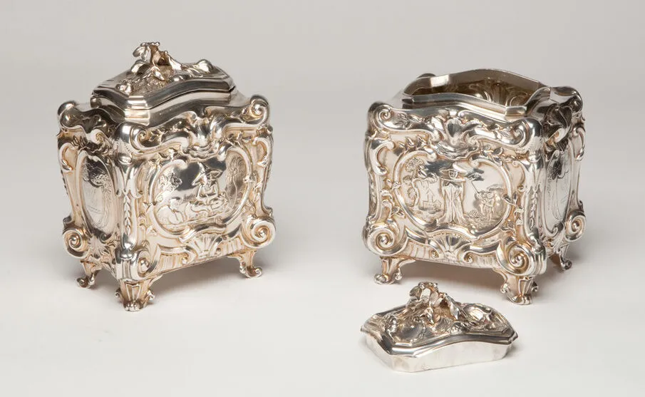 Two silver tea caddies with scenes of tea planters and pickers. Decorated with ornate, rococo scrolls, shells, and flowers, the caddies are opulent and heavily textured, from their four short legs to their tops.