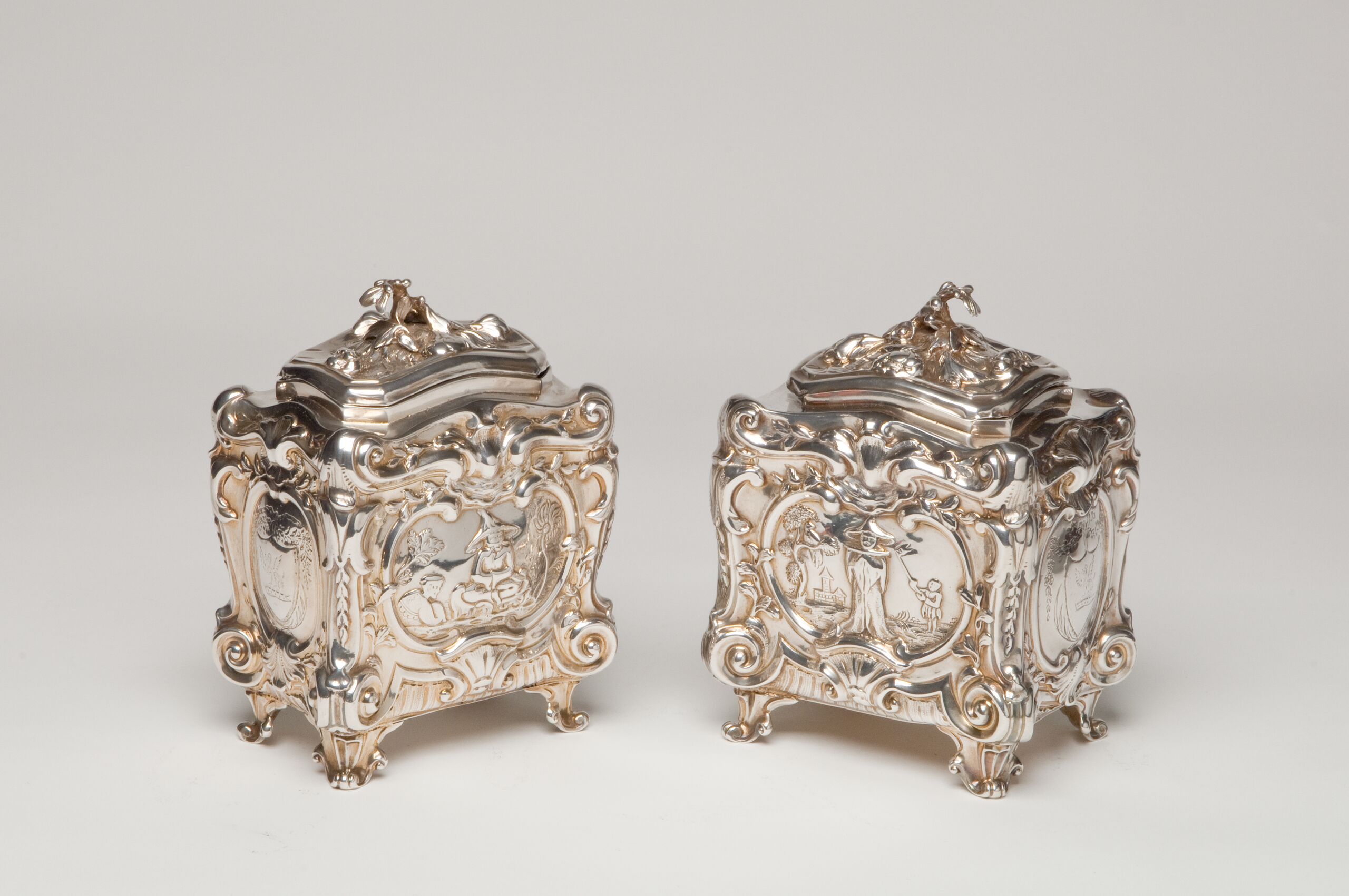 Two silver tea caddies with scenes of tea planters and pickers. Decorated with ornate, rococo scrolls, shells, and flowers, the caddies are opulent and heavily textured, from their four short legs to their tops.
