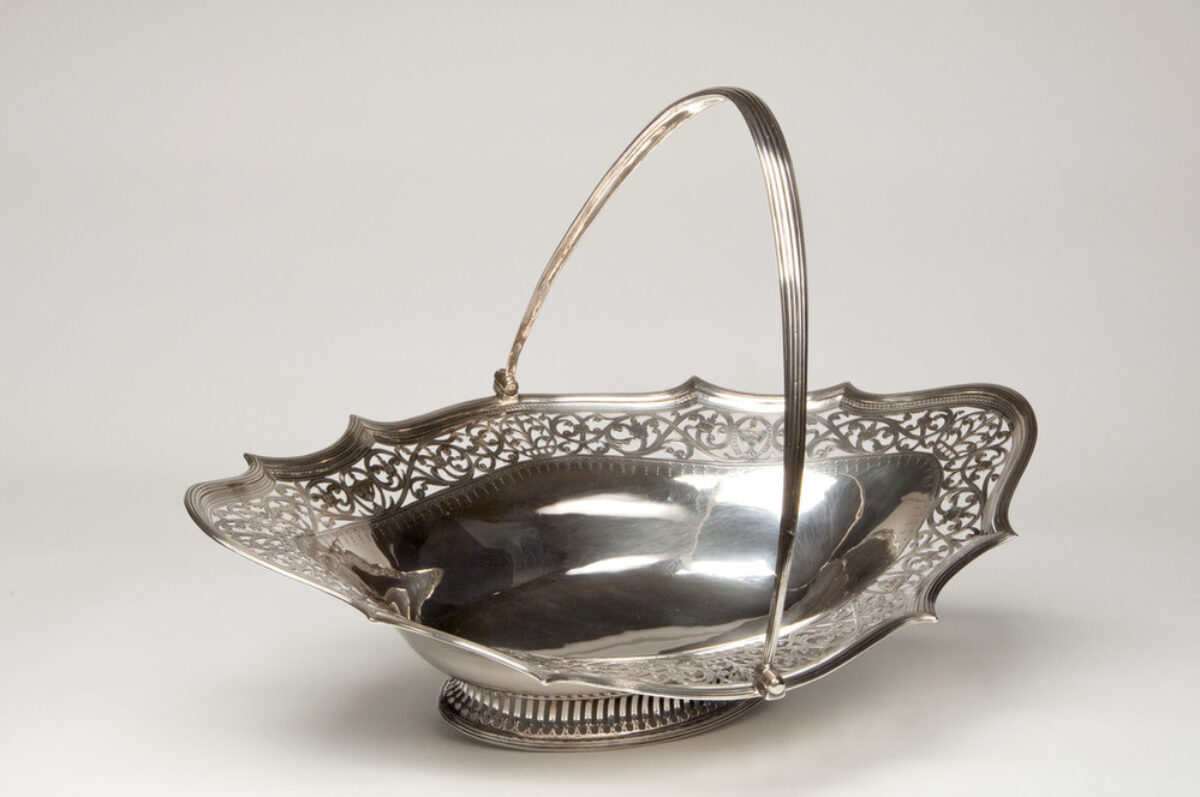 Shallow and oblong, this elegant silver cake basket features a thin, graceful handle, a wide decorative openwork border, and low neoclassical foot.