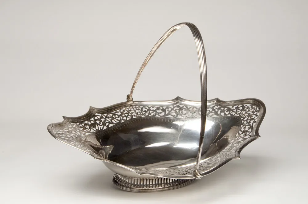 Shallow and oblong, this elegant silver cake basket features a thin, graceful handle, a wide decorative openwork border, and low neoclassical foot.