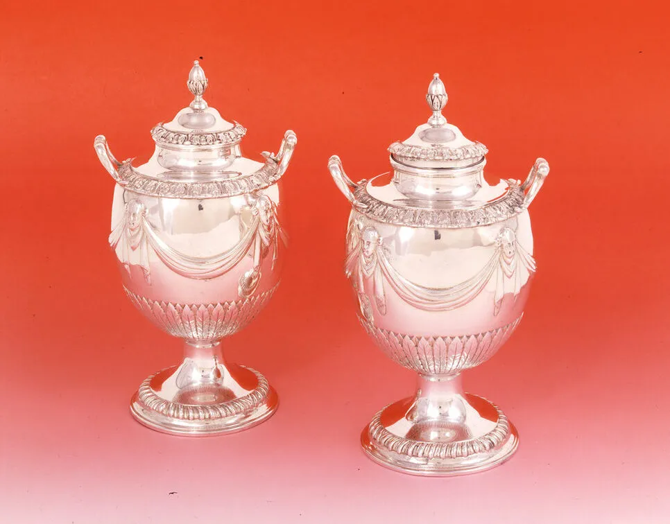 Pair of silver tea caddies with a round base, curved handles and goblet-like shape. Large swaths of polished silver are decorated with drapery punctuated by masks and narrow bands of leaflike patterns. The circular tops are decorated with an oval shape resembling an almond.