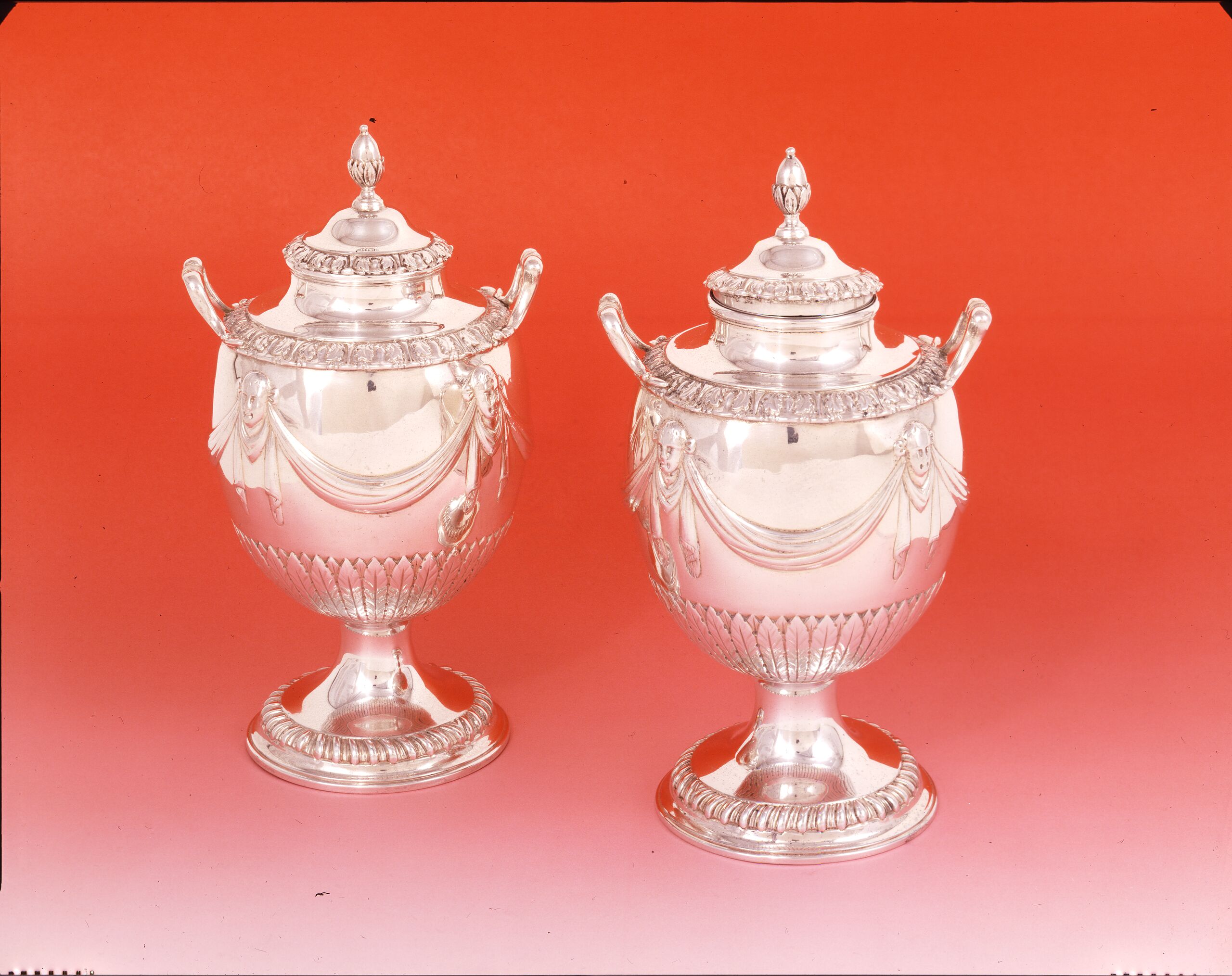 Pair of silver tea caddies with a round base, curved handles and goblet-like shape. Large swaths of polished silver are decorated with drapery punctuated by masks and narrow bands of leaflike patterns. The circular tops are decorated with an oval shape resembling an almond.