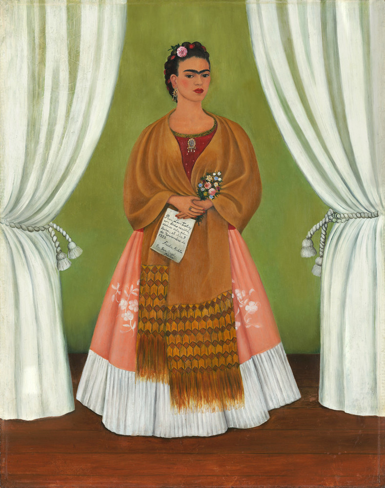 A medium skinned woman stands in a stage-like space framed by white curtains. Beneath black hair woven with red yarn and flowers, heavy brows accent her dark-eyed gaze. Clad in a fringed, honey-toned shawl; long, pink skirt; and gold jewelry, she holds a bouquet and a handwritten letter.