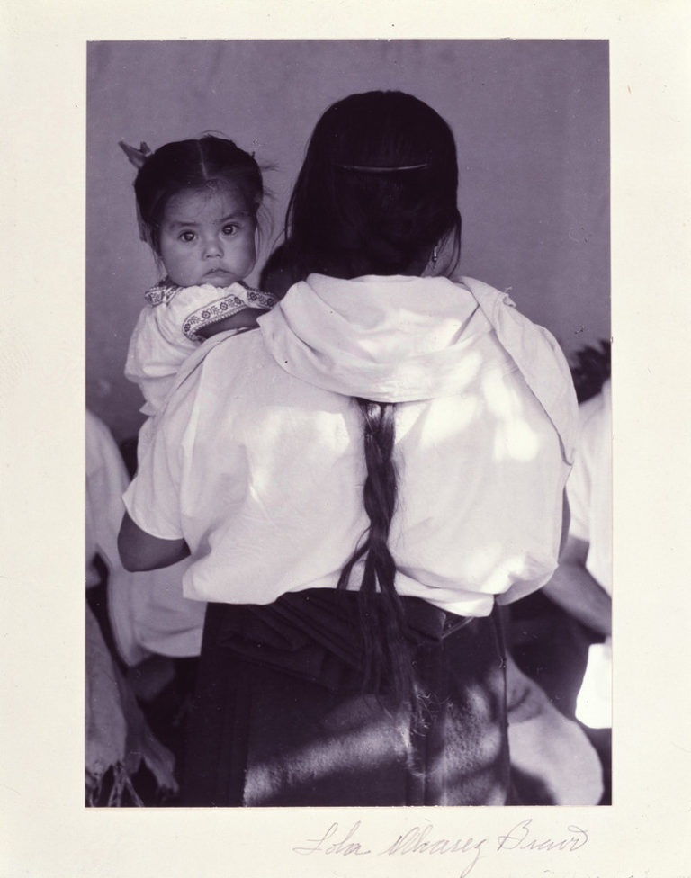 Black-and-white photograph of a woman wearing a dark skirt and light blouse holding a child. The woman faces back and her dark braid reaches past her waist. The dark-haired child stares at the camera and wears a white embroidered dress. Other figures are slightly visible beyond.