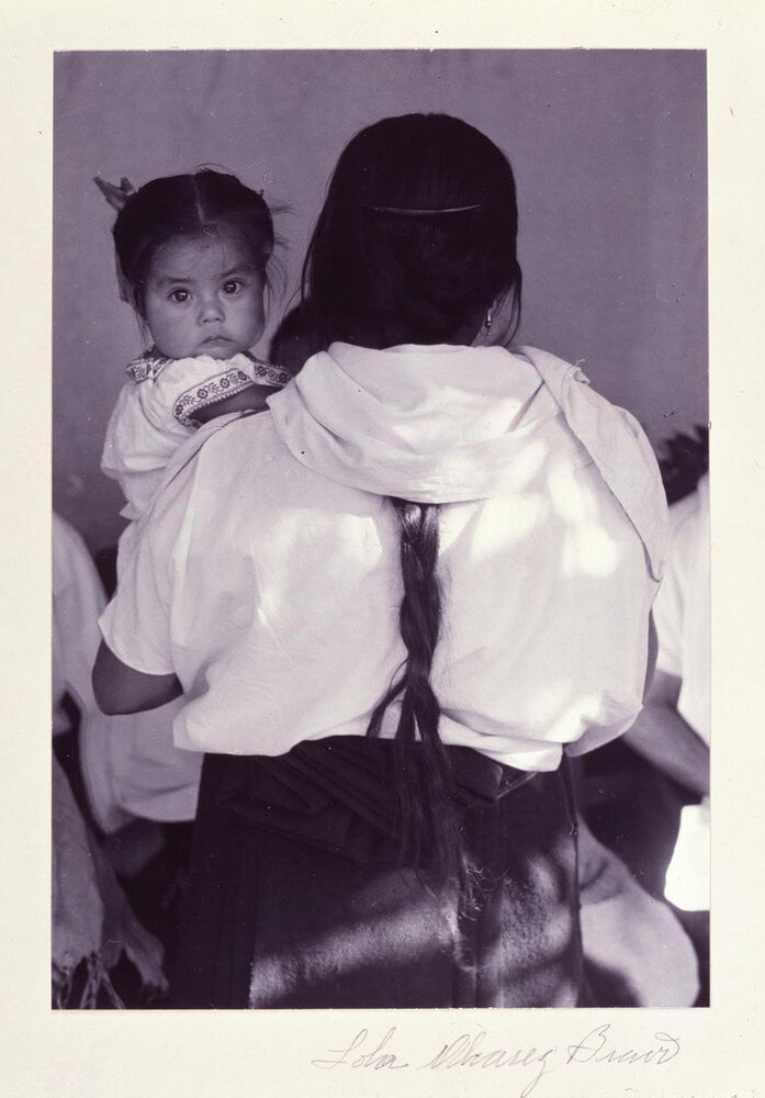 Black-and-white photograph of a woman wearing a dark skirt and light blouse holding a child. The woman faces back and her dark braid reaches past her waist. The dark-haired child stares at the camera and wears a white embroidered dress. Other figures are slightly visible beyond.