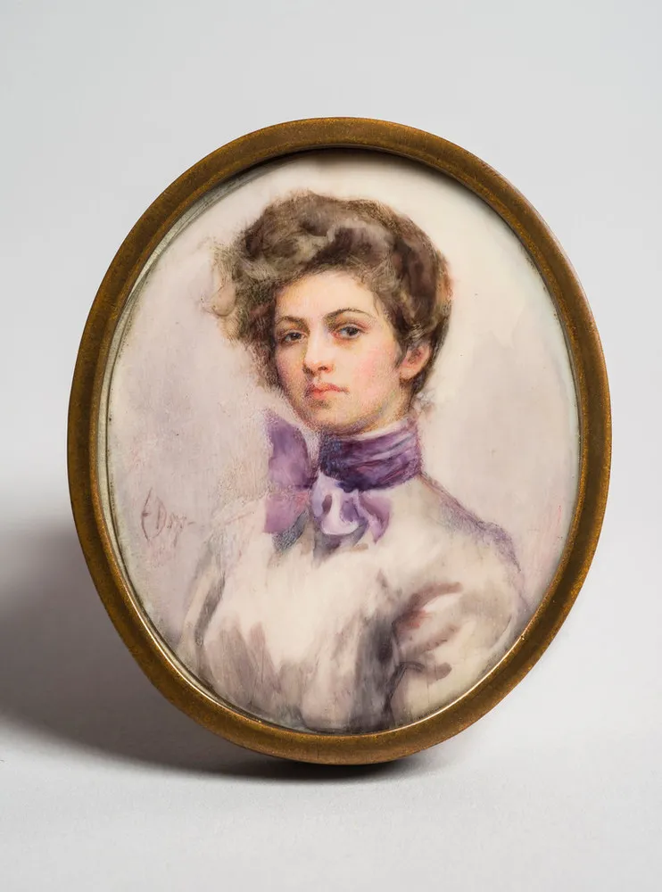 Miniature half-portrait of a light-skinned woman, set in an oval-shaped brass frame, gazing confidently at the viewer. The woman is wearing a Victorian-style white blouse with a purple scarf tied in bow around her neck, her brown hair loosely piled atop her head.