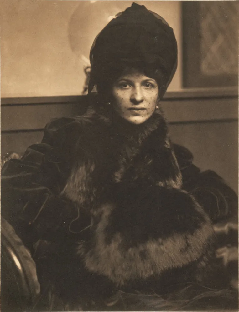 Photographic portrait of a light-skinned adult woman seated and gazing directly at the viewer. She is elegantly dressed in dark clothes and a tall, elaborate bonnet. Rich tonalities pick out the sheen of the fur muff, plush of the velvet coat, and delicate mesh of the veil.