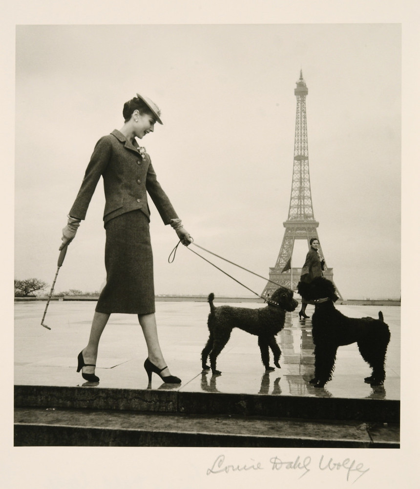 A black-and-white photograph shows a light-skinned, dark-haired woman in a rain-slick plaza with the Eiffel Tower in the distance. Posed in profile, she wears a skirt suit, hat, heels, and gloves. Her left hand holds the leads of two black poodles and her right a furled umbrella.