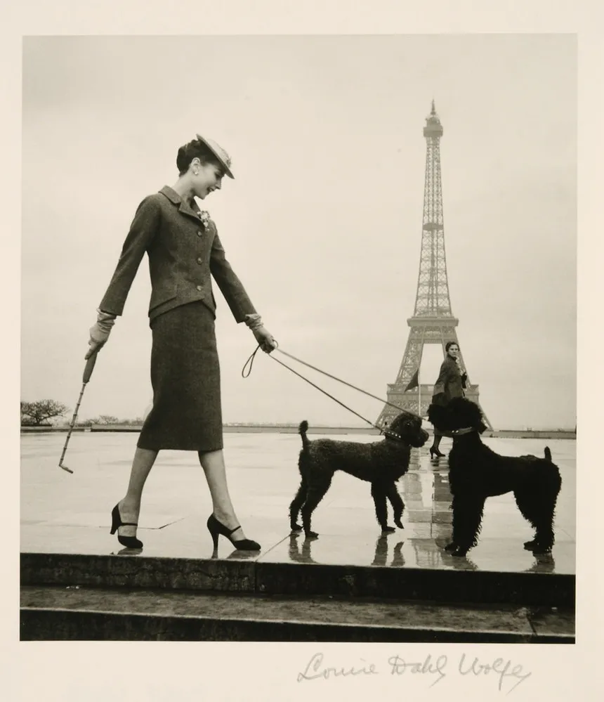 A black-and-white photograph shows a light-skinned, dark-haired woman in a rain-slick plaza with the Eiffel Tower in the distance. Posed in profile, she wears a skirt suit, hat, heels, and gloves. Her left hand holds the leads of two black poodles and her right a furled umbrella.