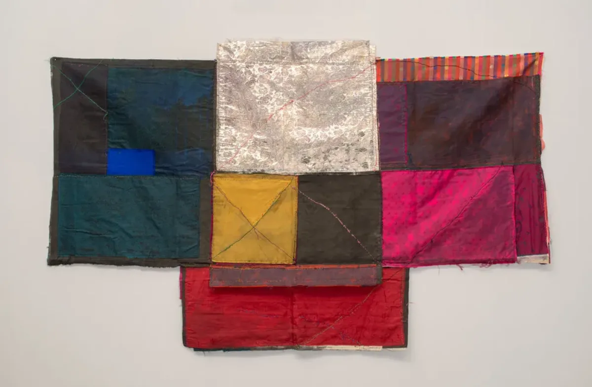 Textile collage of cloth folded and stitched together in loose geometric formations with visible seams making linear patterns. Deep blues on the left and warm colors on the right frame blocks of silver, yellow and grey in the center, which extends into a slightly longer base.