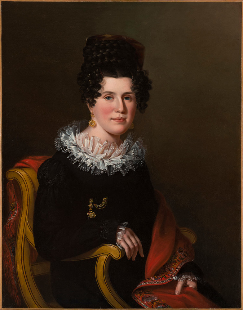 A light-skinned woman with dark curly hair tied up on her head is sitting on a wooden chair. Her right hand is rested on the armrest. She is wearing a black dress with a white transparent collar.