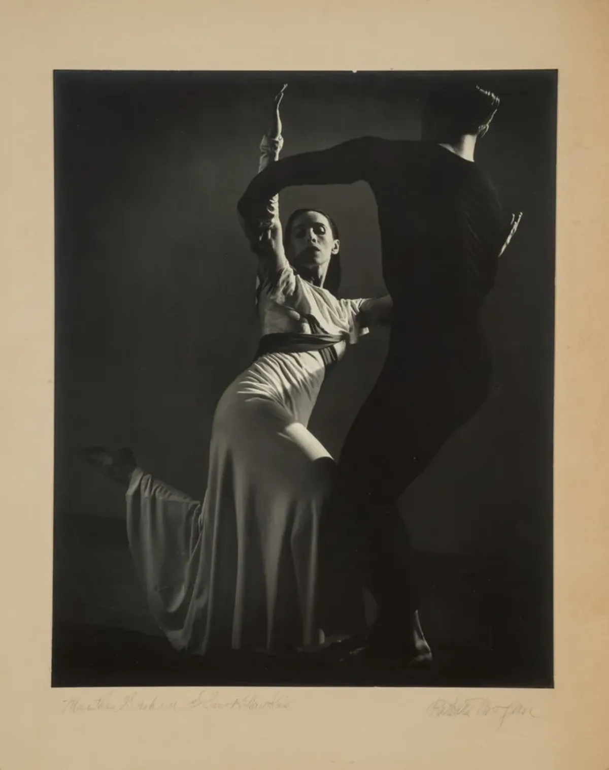 Black-and-white photograph of a man, dressed in black, and Martha Graham, in white, dancing. Graham, lit from above, strikes one of her famous angular poses, both legs bent, the back foot pointing behind her. Her raised arms are held by the man, who stands with his back to us.