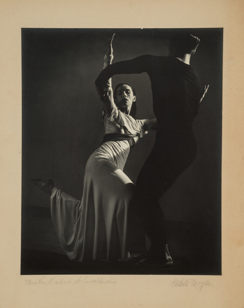 Black-and-white photograph of a man, dressed in black, and Martha Graham, in white, dancing. Graham, lit from above, strikes one of her famous angular poses, both legs bent, the back foot pointing behind her. Her raised arms are held by the man, who stands with his back to us.