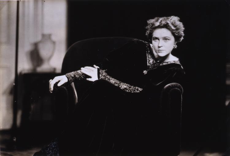 Black and white photo of Eva Le Gallienne seated on a dark chair, her head shifts left. Dark tones dominate the photograph, drawing focus to her illuminated face and hands, which are balanced by a glimpse of the room behind her. Her arms draped in ornately decorated sleeves, reach right.