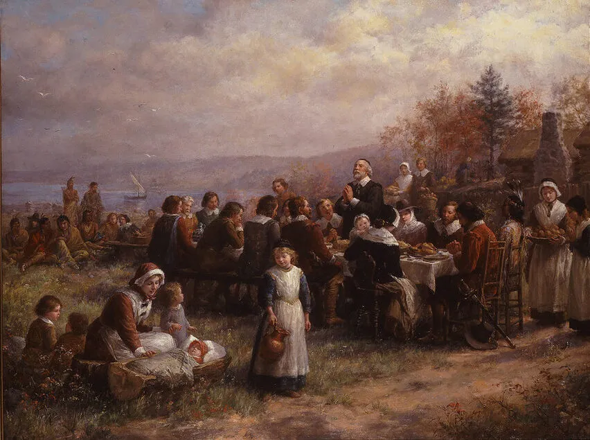 A detailed painting portrays an idealized version of the “First Thanksgiving.” At right, austerely garbed men and women and one Native American sit at a long table as an old man leads a prayer. A woman attends small children at the left, and group of Native Americans sits nearby.