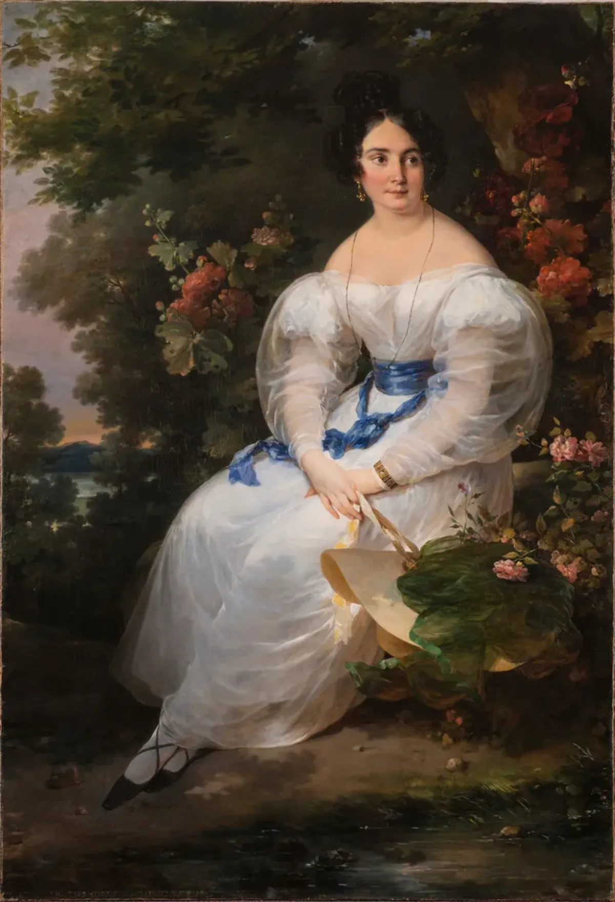 Full-length portrait of a light-skinned woman with black hair pinned up, gazing off to the viewer's right. She wears a delicate white gown with billowing sleeves and blue silk sash and holds a straw sun hat in front of a large dark tree surrounded by flowering bushes.