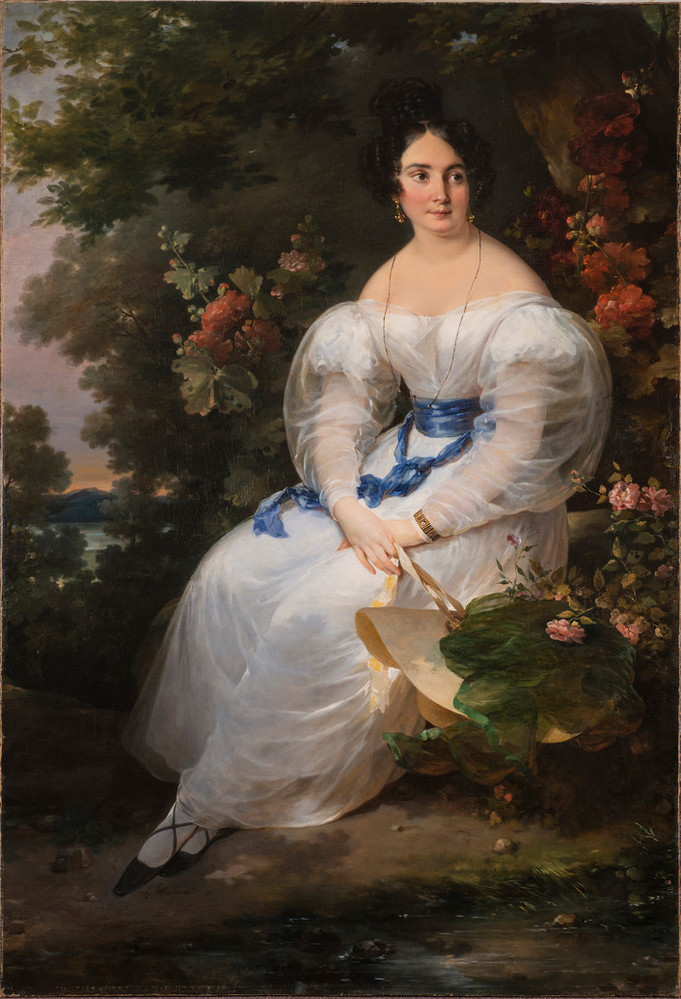 Full-length portrait of a light-skinned woman with black hair pinned up, gazing off to the viewer's right. She wears a delicate white gown with billowing sleeves and blue silk sash and holds a straw sun hat in front of a large dark tree surrounded by flowering bushes.