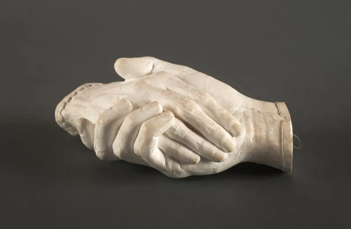 Life-sized plaster cast sculpture of a man's hand on the bottom holding a woman's hand on top. The matte white surface reveals the textures of the skin and fingernails. The man's hand is capped with a plain thin cuff, while the woman's is capped with a delicate lace cuff.