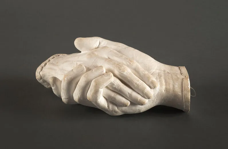 Life-sized plaster cast sculpture of a man's hand on the bottom holding a woman's hand on top. The matte white surface reveals the textures of the skin and fingernails. The man's hand is capped with a plain thin cuff, while the woman's is capped with a delicate lace cuff.