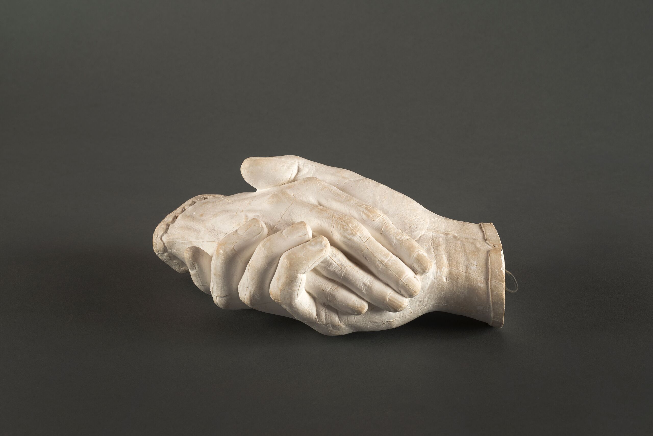 Life-sized plaster cast sculpture of a man's hand on the bottom holding a woman's hand on top. The matte white surface reveals the textures of the skin and fingernails. The man's hand is capped with a plain thin cuff, while the woman's is capped with a delicate lace cuff.