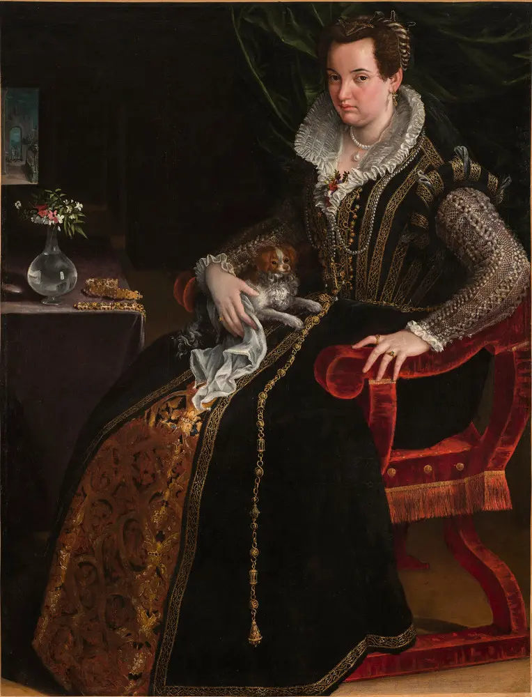 A light-skinned, dark-haired woman wearing pearl and gold jewelry and a black-velvet, lace, and brocade gown poses with her lap dog. Seated in a red velvet chair in the foreground, she makes eye contact with viewers while pressing her hand on the armrest, as if to stand.