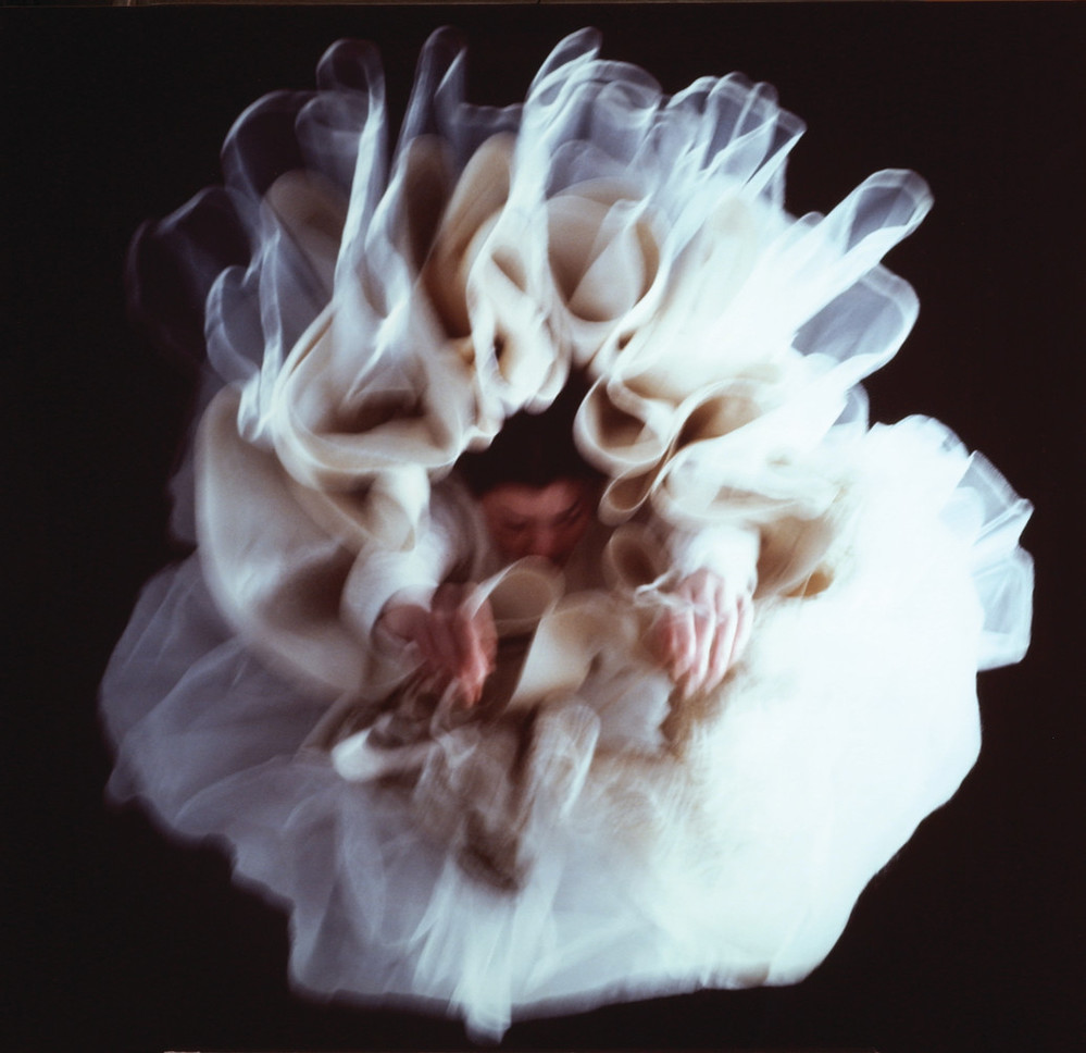 Large, glossy, blurry, color photograph of a light-skinned woman hanging upside-down directly above the viewer. Her arms hang loosely above her head, enveloped in diaphanous white skirts billowing around her, set against a black background, creating a flower-like appearance.