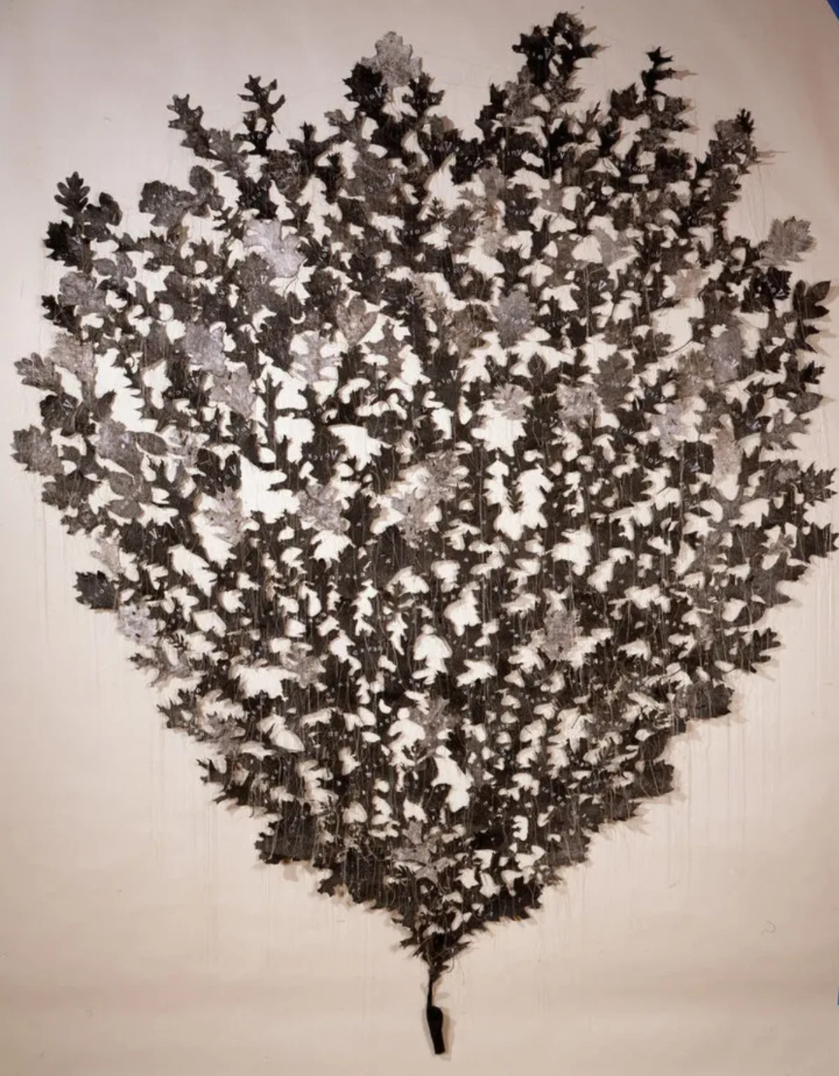 Hundreds of light- and dark-gray shapes resembling oak leaves are mounted directly to a wall in the shape of an ice-cream cone. They appear to sprout from the index finger of a small, coal-black hand mounted near the floor. Delicate strands of thread hang down from the leaves.