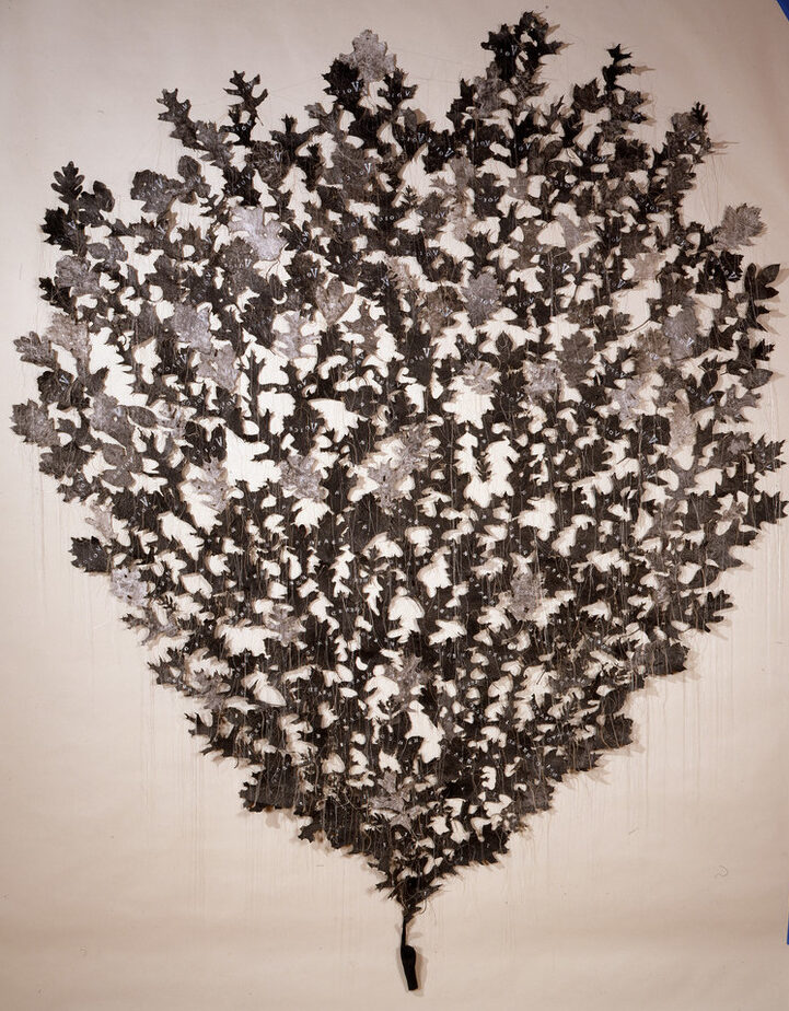 Hundreds of light- and dark-gray shapes resembling oak leaves are mounted directly to a wall in the shape of an ice-cream cone. They appear to sprout from the index finger of a small, coal-black hand mounted near the floor. Delicate strands of thread hang down from the leaves.