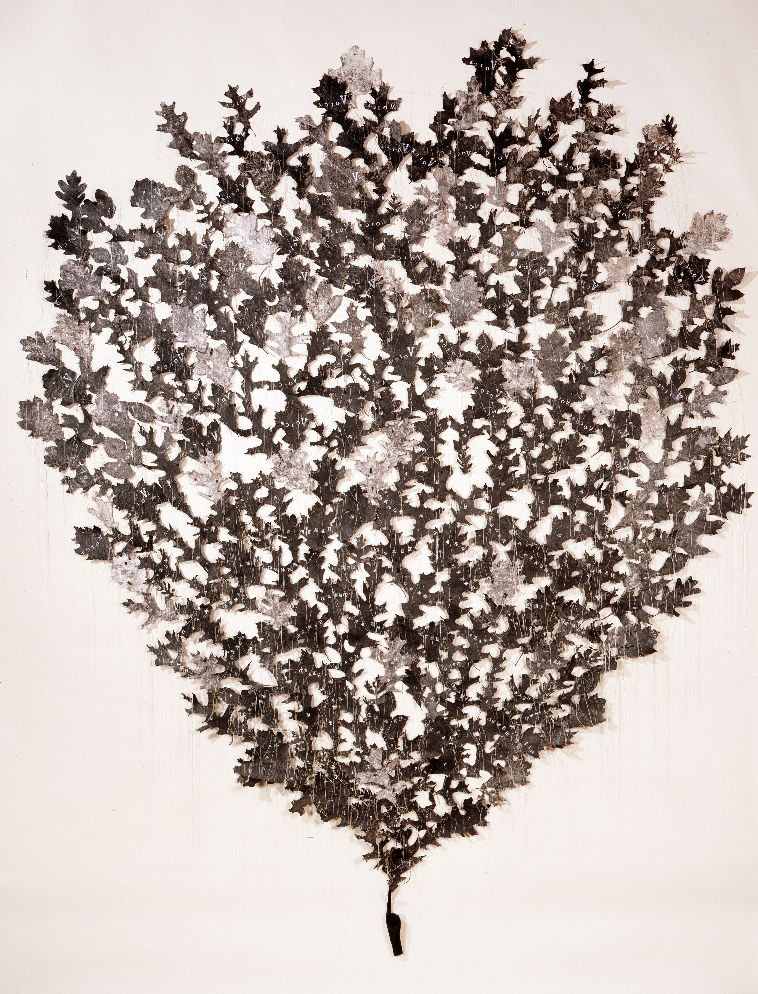 Numerous light- and dark-gray shapes resembling oak leaves are mounted directly to a wall in the shape of an ice-cream cone. They appear to sprout from the index finger of a small, coal-black hand mounted near the floor. Delicate strands of thread hang down from the leaves.
