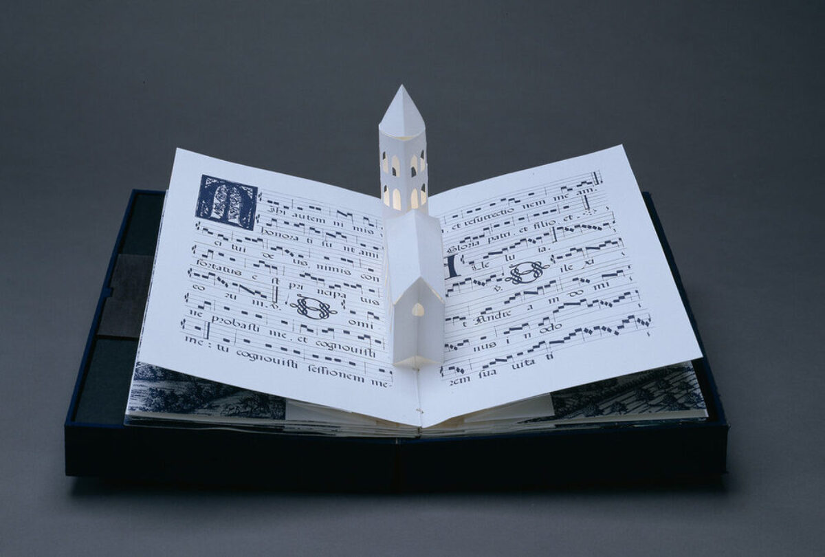 Bound in a dark blue fabric-covered binding, the book opens to display five pop-up towers illuminated from within by a tiny bulb. The featured page includes a white country church with a steeple, set into pages featuring musical scores presented in manuscript style.