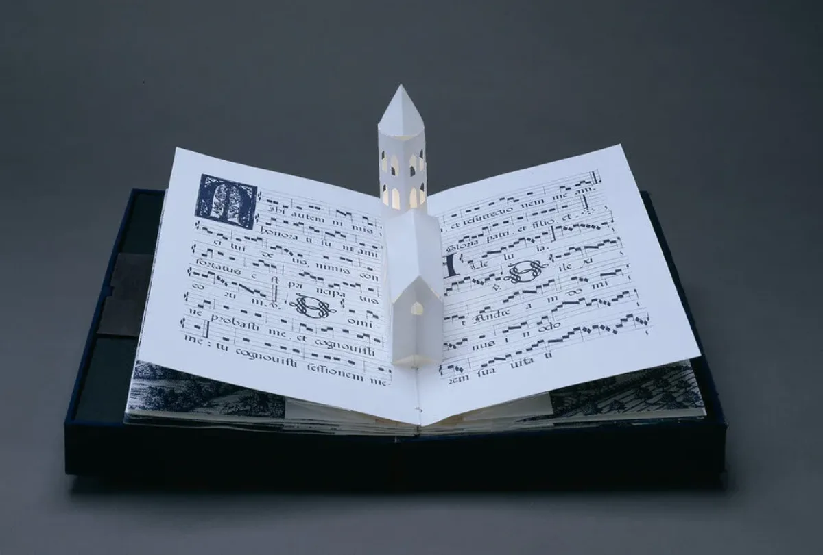 Bound in a dark blue fabric-covered binding, the book opens to display five pop-up towers illuminated from within by a tiny bulb. The featured page includes a white country church with a steeple, set into pages featuring musical scores presented in manuscript style.