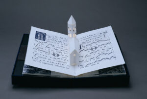 Bound in a dark blue fabric-covered binding, the book opens to display five pop-up towers illuminated from within by a tiny bulb. The featured page includes a white country church with a steeple, set into pages featuring musical scores presented in manuscript style.