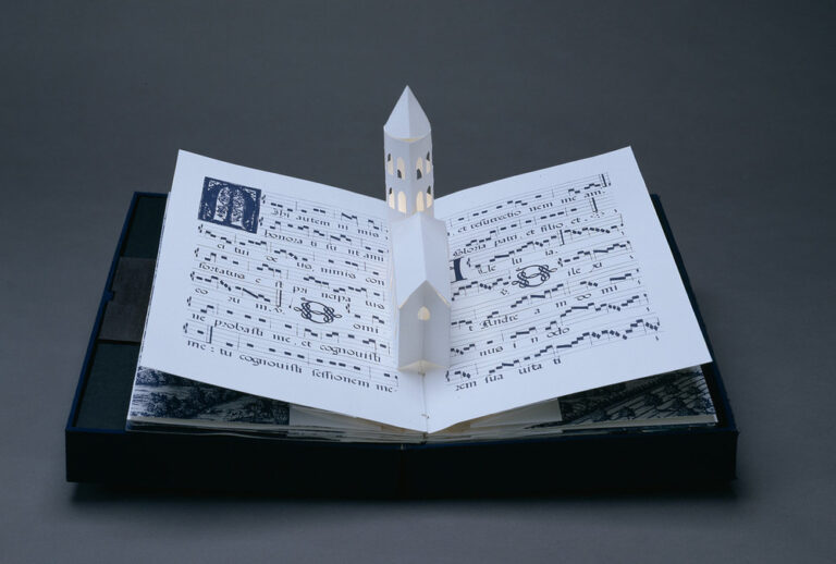 Bound in a dark blue fabric-covered binding, the book opens to display five pop-up towers illuminated from within by a tiny bulb. The featured page includes a white country church with a steeple, set into pages featuring musical scores presented in manuscript style.