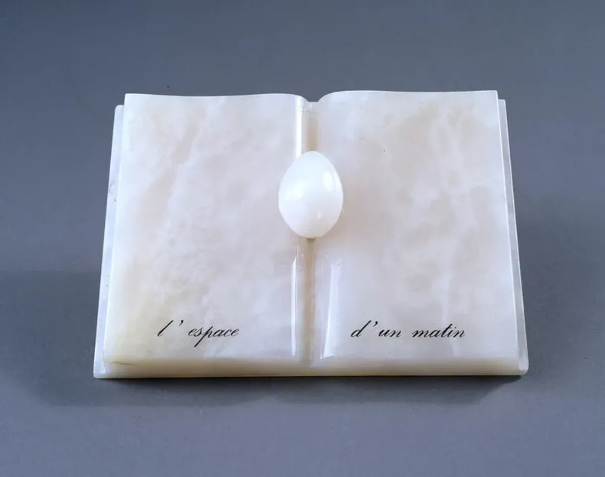 An open book with an egg nestled in the spine, all made of rose-colored onyx. An inscription in French painted in black at the bottom reads, "L’espace d’un matin" (The span of a morning).