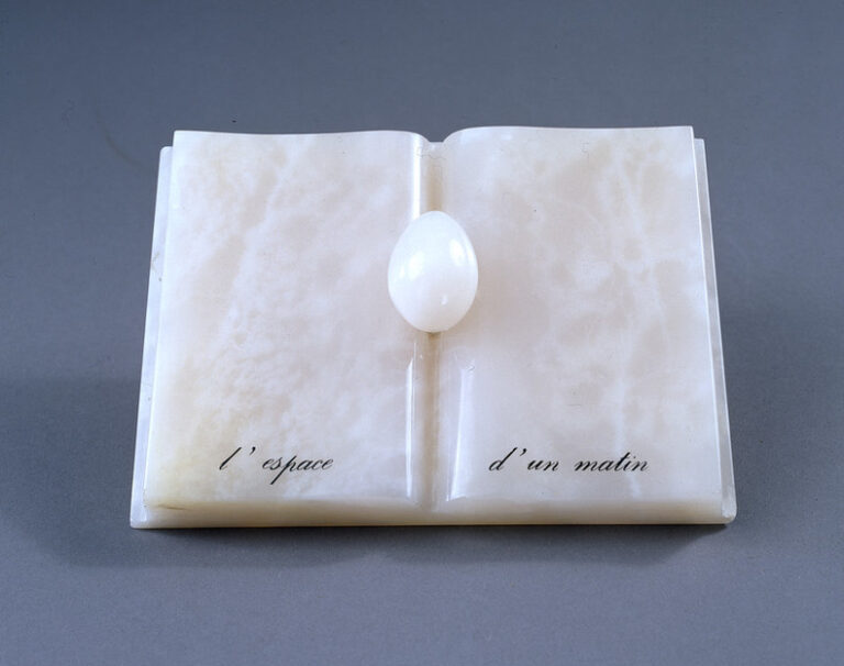 An open book with an egg nestled in the spine, all made of rose-colored onyx. An inscription in French painted in black at the bottom reads,