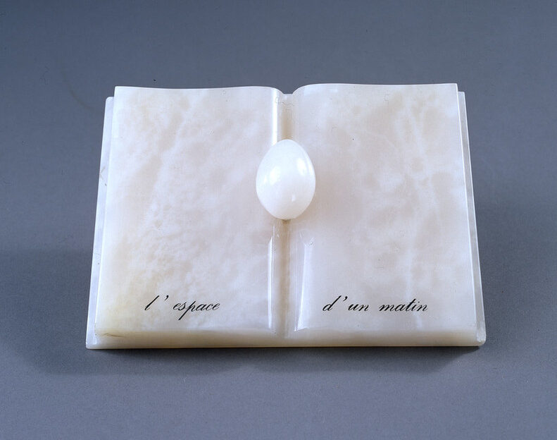 An open book with an egg nestled in the spine, all made of rose-colored onyx. An inscription in French painted in black at the bottom reads, 