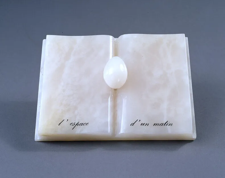 An open book with an egg nestled in the spine, all made of rose-colored onyx. An inscription in French painted in black at the bottom reads, "L’espace d’un matin" (The span of a morning).