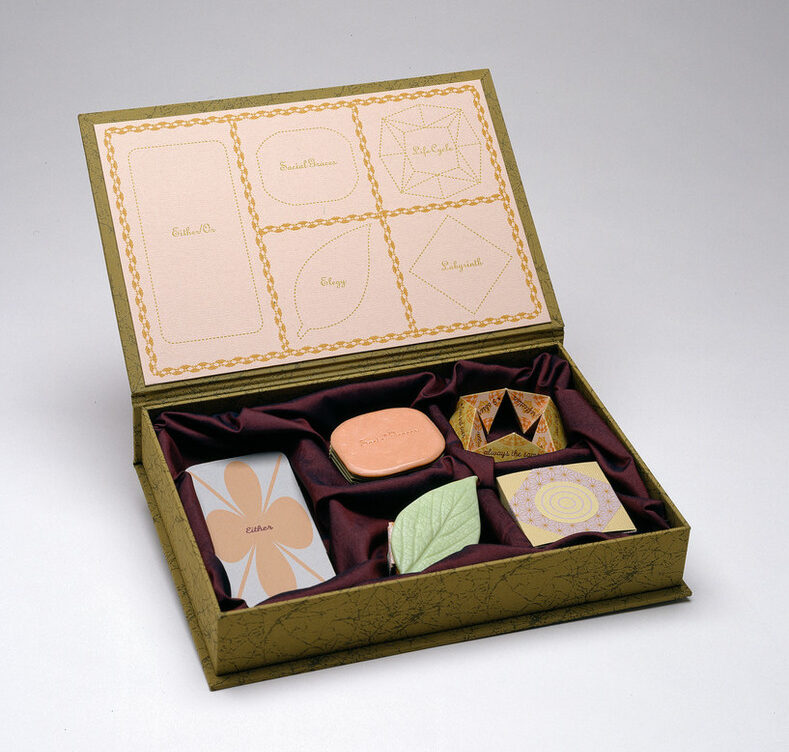 A green-gold box with its attached lid flipped open holds five objects shaped liked candies resting on burgundy fabric. The inside of the lid displays outlines of each object, with the words "life cycle," "labyrinth," "elegy," "either/or," and "social graces" inside the outlines.