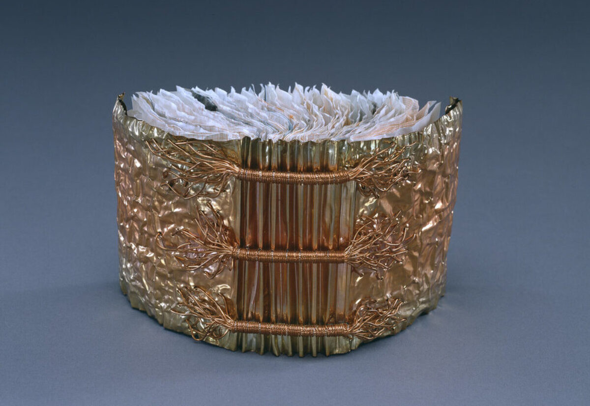 A book whose cover is made of copper is open and standing upright with its spine facing viewer. Three horizontal bands of wound copper wire extend the width of the accordion pleated spine. Crumpled paper comprises the pages of the book.