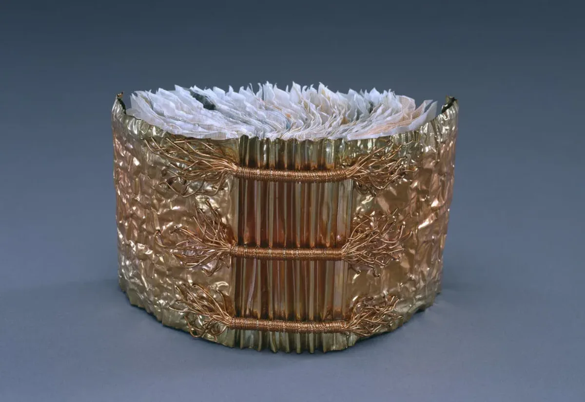 A book whose cover is made of copper is open and standing upright with its spine facing viewer. Three horizontal bands of wound copper wire extend the width of the accordion pleated spine. Crumpled paper comprises the pages of the book.