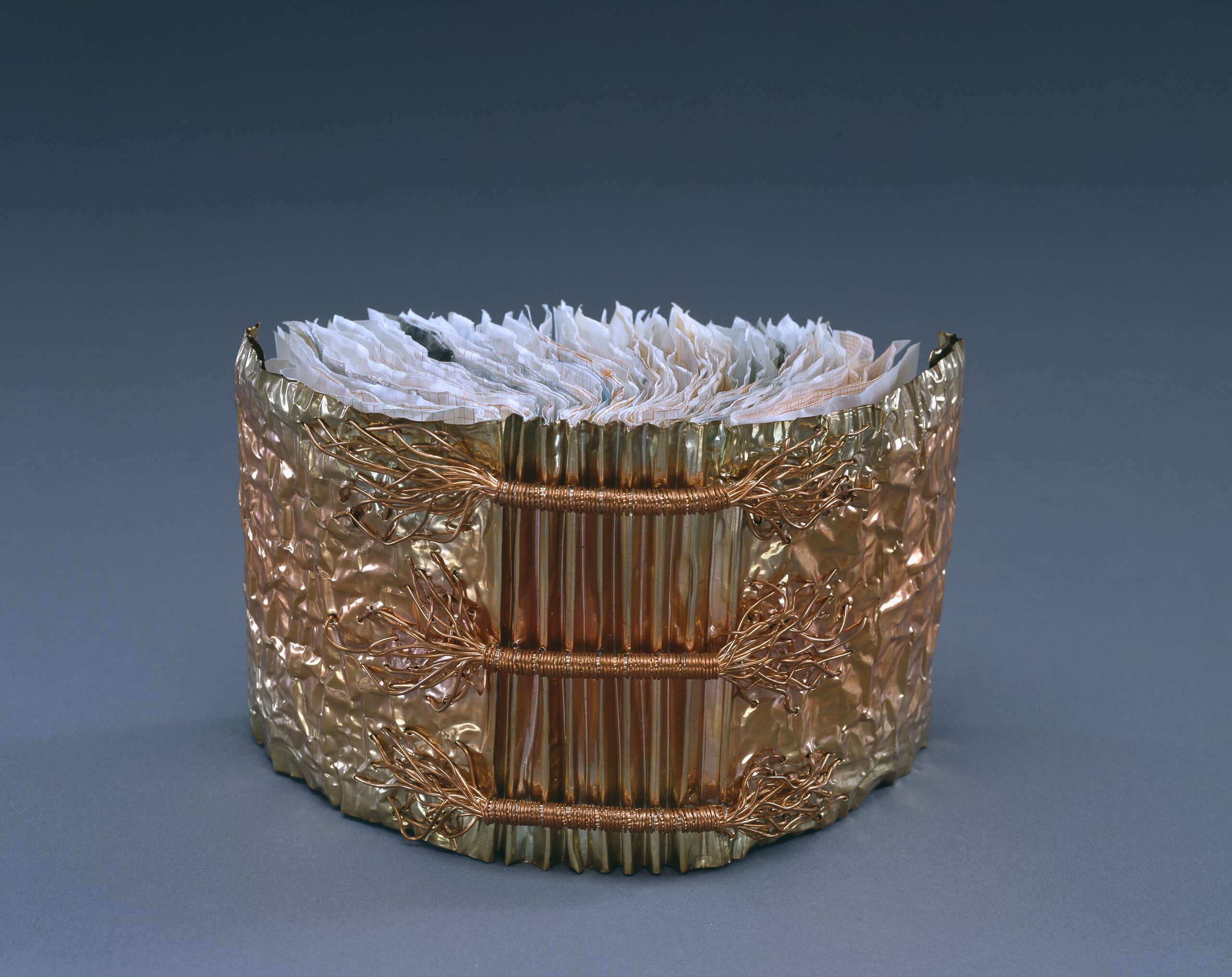 A book whose cover is made of copper is open and standing upright with its spine facing viewer. Three horizontal bands of wound copper wire extend the width of the accordion pleated spine. Crumpled paper comprises the pages of the book.