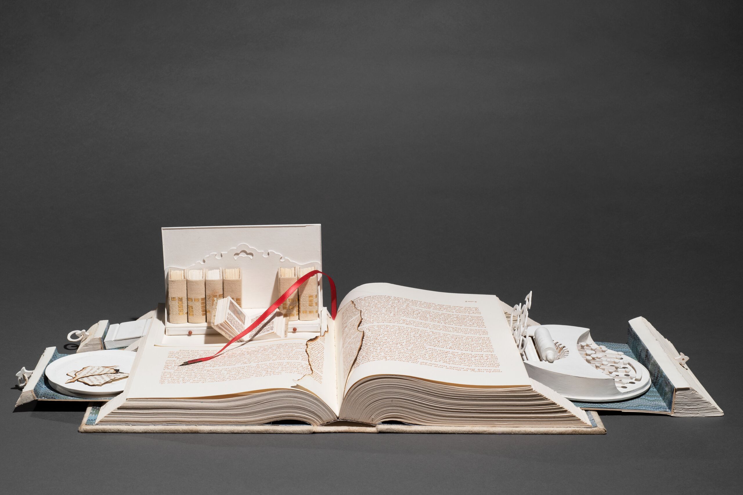 A white book opens like a Swiss army knife; its pages cut to create a sculpture. Opened to the center, pull-out drawers open from the sides, filled with paper-made objects. On the right, a typewriter-like object. On the left, a box with smaller books, including one that is also open.