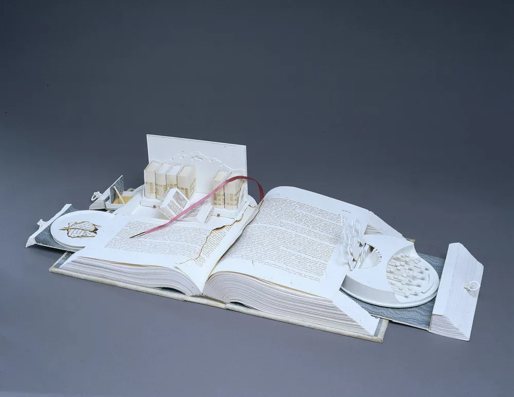 A white book opens like a Swiss army knife; its pages cut to create a sculpture. Opened to the center, pull-out drawers open from the sides, filled with paper-made objects. On the right, a typewriter-like object. On the left, a box with smaller books, including one that is also open.