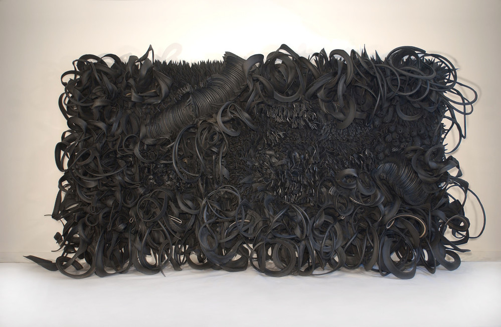 A large-scale, horizontal sculpture consists of black rubber tires and tubing that has been sliced, stripped, woven, looped, twisted and otherwise manipulated into an expressive and abstract high-relief tableau.