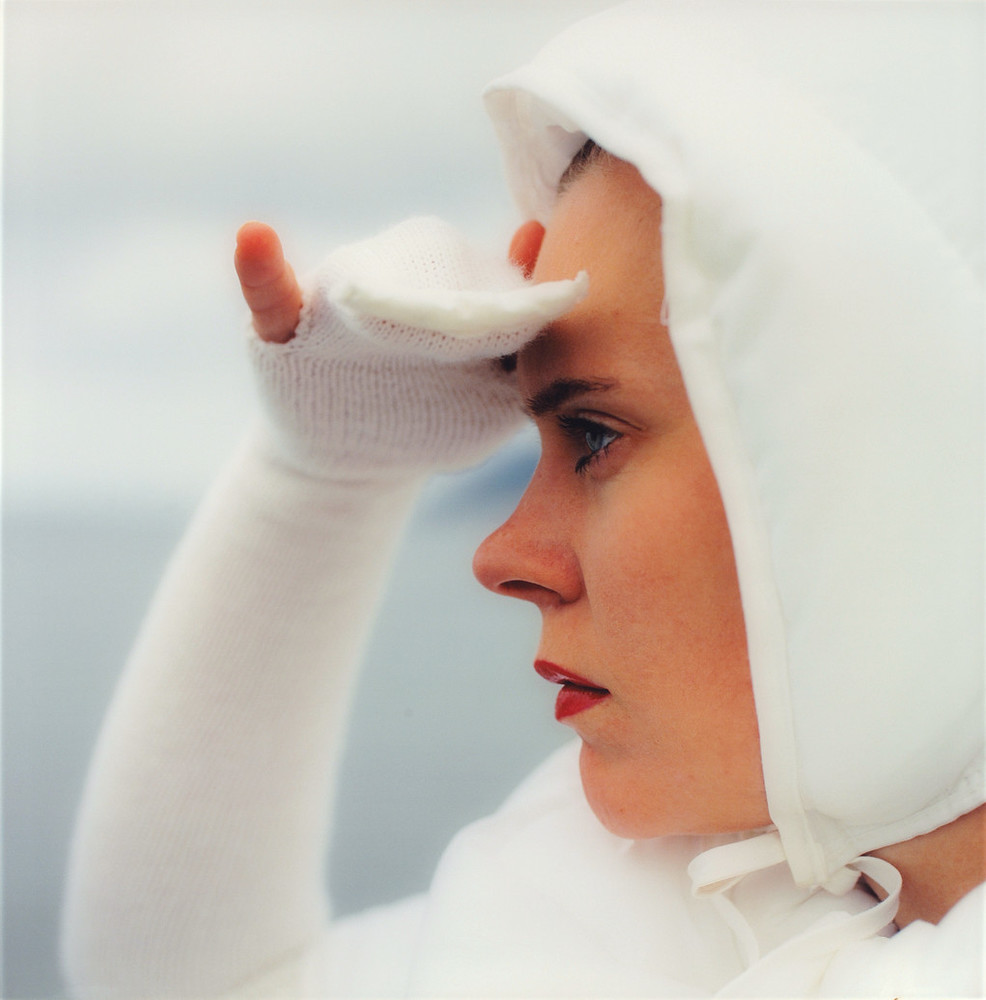 Close-up of a light-skinned woman in profile looking to the left. She is wearing all white including a bonnet and she is holding her hand up as if to shield her eyes from the sun.