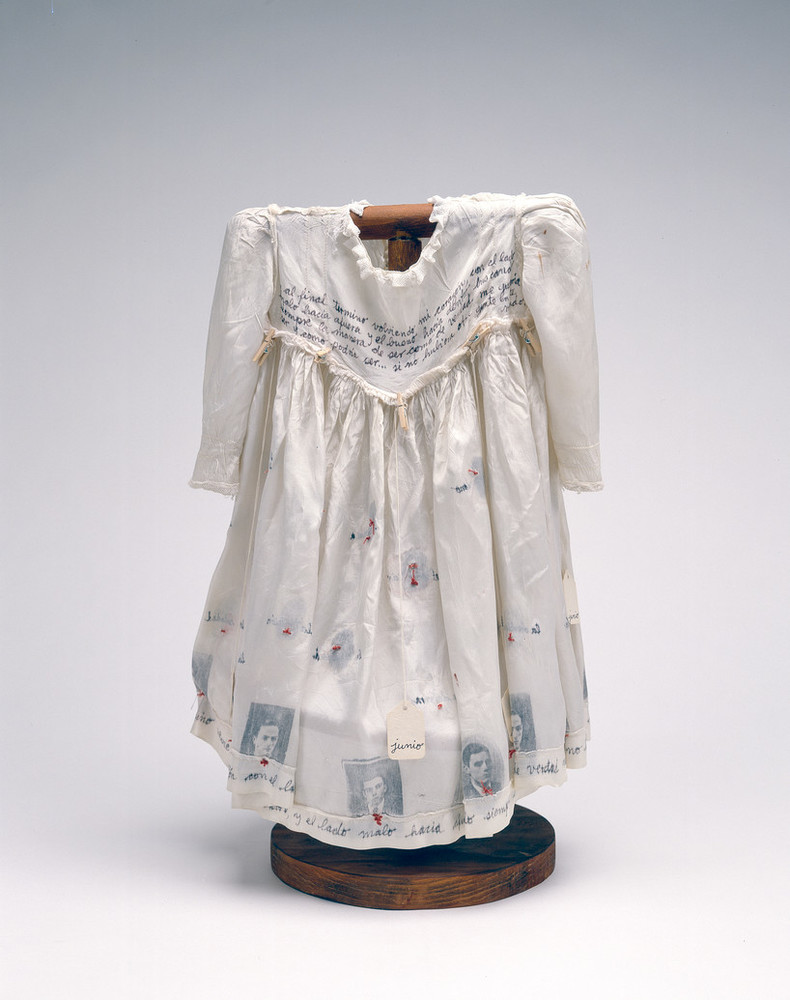 Hung on a dark wood, T-shaped form, a child's ivory-colored silk dress serves as a canvas for photographs, calligraphy, stitching, and the words of Anne Frank.