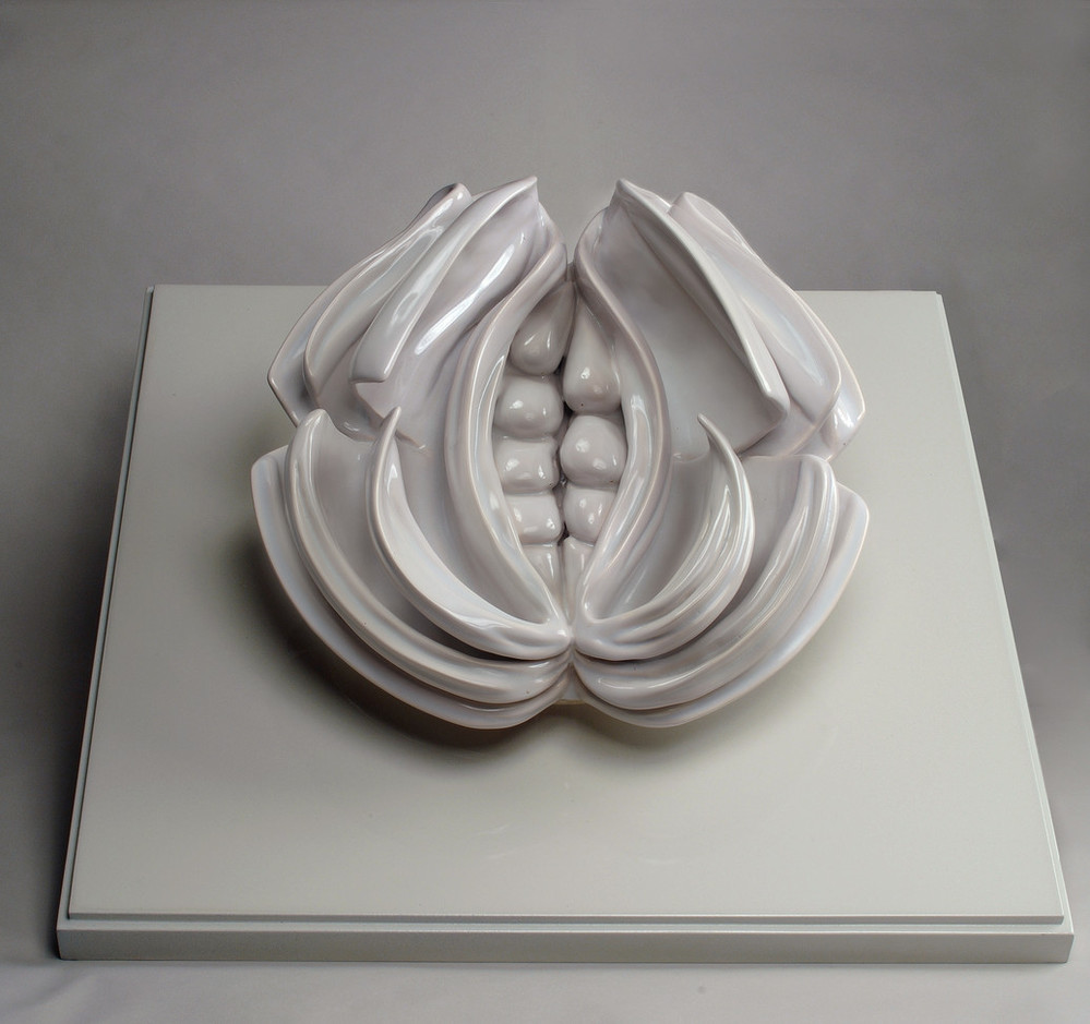 A glossy white porcelain sculpture rests on a flat white slab. The sculpture is shaped like a vulva with a core of seed-like shapes surrounded by three-dimensional petals that curl at the edges, mimicking pages of an open book.