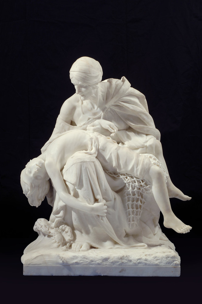 Large sculpture in white marble depicts a seated older woman cradling the body of a young boy whose limp body falls over her legs. The boy's legs are tangled in a fisherman's net while waves crash at the woman's feet.