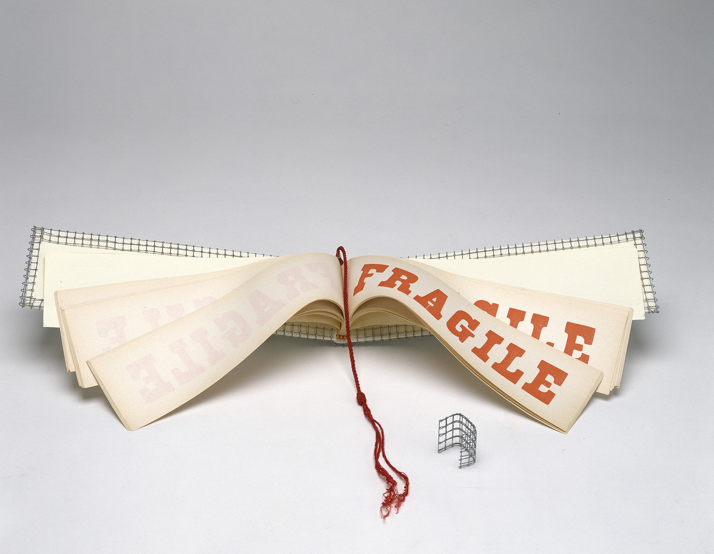 A wide and narrow book opened to display pages with the word FRAGILE in red block letters. A red tassel hangs down the center of the book and the cover is wrapped with wire mesh.